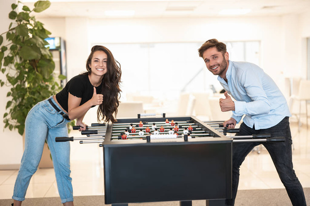 two young colleagues brunette beautiful woman and handsome man playing table football during break at the office showing like signs - Foto, afbeelding