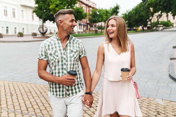 Image of cheerful young couple holding hands together while walking through city street with paper cups - Фото, изображение