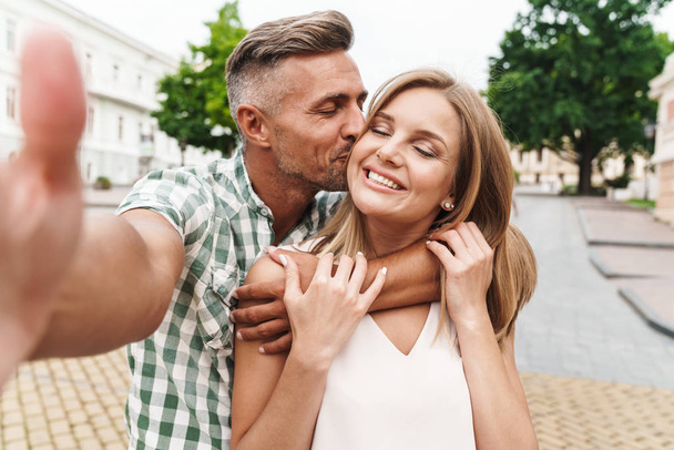 Image of loving young couple kissing and taking selfie photo together while walking through city street - Photo, image