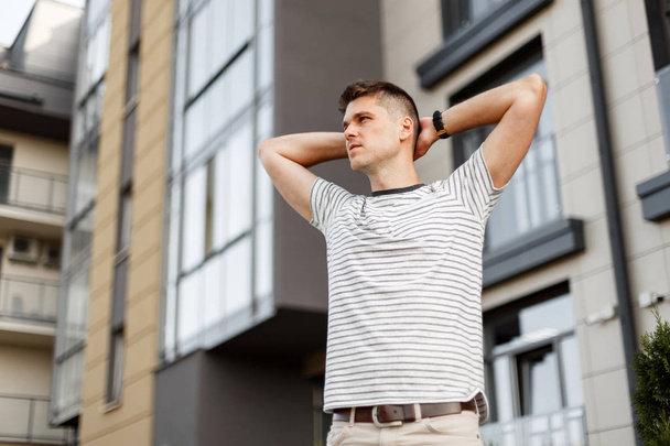 Pohledný mladý muž s módním účesu v módní proužkované košili, který se nachází ve městě nedaleko budovy. Pohledný model chodí po ulici v letním dnu. Oblečení módního muže. - Fotografie, Obrázek