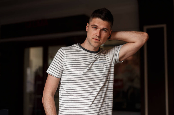 Handsome young man model with stylish hairstyle with beautiful gray eyes in a fashionable striped t-shirt posing standing in a dark room. Attractive guy. Trendy men's summer clothing. - Foto, Imagem