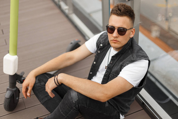 Elegante joven hipster hombre fresco en gafas de sol oscuras en ropa de verano de moda descansando después de montar una scooter eléctrico cerca de una pared de vidrio en la ciudad. Guy se está relajando en la terraza de un café de verano
. - Foto, Imagen