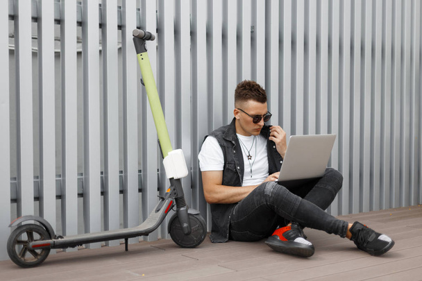 Urban jeune homme hipster dans un gilet tendance en jean vintage en baskets en lunettes de soleil utilise un ordinateur portable moderne près d'un mur en métal. Beau gars se reposer après avoir roulé un scooter électrique sur une journée d'été
. - Photo, image