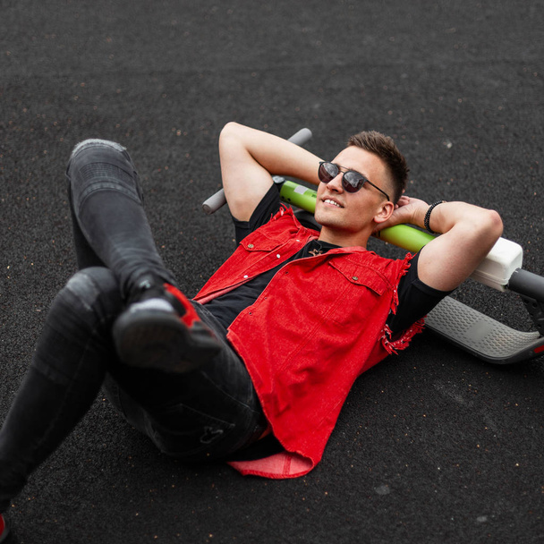 Das fröhliche junge Männermodel in modischer Jeansweste, schwarzer Jeans und Sonnenbrille genießt es, auf dem Bürgersteig zu liegen. amerikanischer Typ Hipster mit einem süßen Lächeln entspannt sich nach der Fahrt auf einem Elektroroller. - Foto, Bild