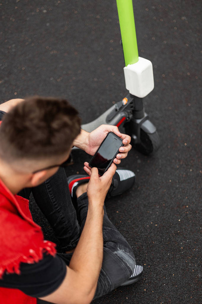 Κορυφαία άποψη ενός νεαρού άνδρα με ένα μοντέρνο χτένισμα σε ένα μοντέρνο κόκκινο γιλέκο denim με ένα σκούτερ και με ένα τηλέφωνο στα χέρια. Κομψός σύγχρονος τύπος αναπαύεται στο πεζοδρόμιο σε εξωτερικούς χώρους. Καλοκαίρι. - Φωτογραφία, εικόνα