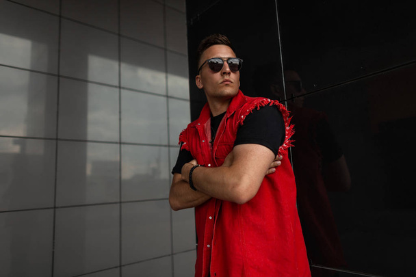 Homem hipster atraente jovem em óculos de sol elegantes em uma camiseta em um colete de ganga vermelho na moda posa perto de uma parede vintage preta na cidade. Bonito modelo moderno. Roupas de verão masculinas na moda
. - Foto, Imagem