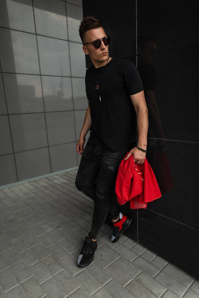 O jovem bonito na moda em roupas pretas de verão na moda com uma jaqueta de ganga vermelha em óculos escuros em tênis está descansando perto de uma parede moderna na cidade. Um americano. Roupas masculinas elegantes
. - Foto, Imagem