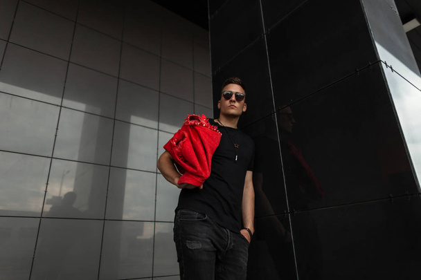 Muodikas hipster nuori mies punainen denim liivi musta T-paita farkut aurinkolasit poseeraa lähellä seinää kadulla kaupungissa. Komea tyyppi malli ulkona. Nuorten katumuoti
. - Valokuva, kuva