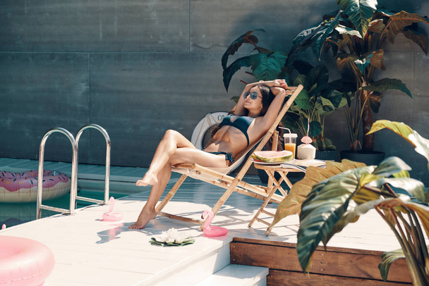 Havuz başında açık havada güneşlenirken bikinili çekici genç kadın dinlendirici ve gülümseyen    - Fotoğraf, Görsel