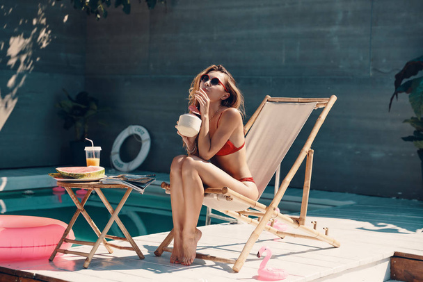 屋外のプールサイドで日光浴をしながらリラックスして爽やかなカクテルを楽しむビキニの魅力的な若い女性 - 写真・画像