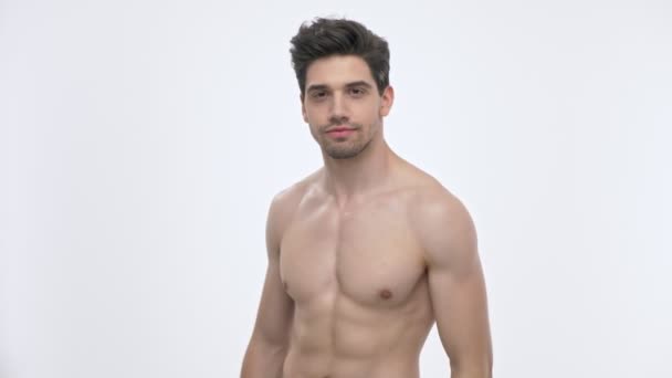 Zijaanzicht van zelfverzekerde jonge brunette man met naakte torso toont zijn biceps met verhoogde arm terwijl het kijken naar de camera over witte achtergrond geïsoleerd - Video
