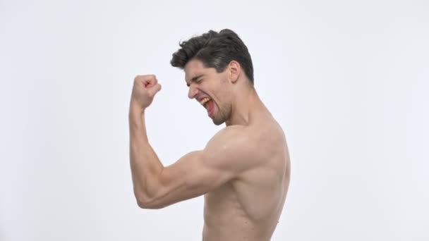 Side άποψη του χαρούμενο νεαρό μελαχρινή άντρα με γυμνό κορμό ουρλιάζοντας και δείχνοντας νικητή χειρονομία με το χέρι, ενώ κοιτάζοντας την κάμερα πάνω από λευκό φόντο απομονωμένη - Πλάνα, βίντεο