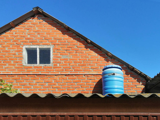 barril azul. en el techo contra el cielo azul y el ladrillo rojo. día soleado. mejora de una casa privada. recipiente de plástico para calentar agua, ducha exterior
 - Foto, Imagen