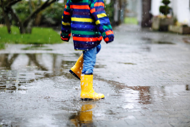 Close-up of kid wearing yellow rain boots and walking during sleet, rain and snow on cold day. Child in colorful fashion casual clothes jumping in a puddle. Having fun outdoors - Photo, Image