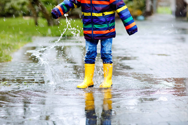 Κοντινό πλάνο του παιδιού φορώντας κίτρινες μπότες βροχής και περπατώντας κατά τη διάρκεια του ύπνου, της βροχής και του χιονιού την κρύα μέρα. Παιδί σε πολύχρωμο μόδα casual ρούχα άλμα σε μια λακκούβα. Διασκεδάζοντας σε εξωτερικούς χώρους - Φωτογραφία, εικόνα