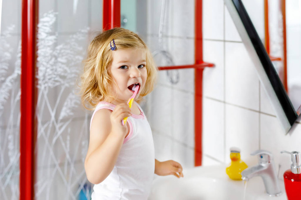 Cute adorable toddler girl holding toothbrush and brushing first teeth in bathroom after sleeping. Gorgeous baby child learning to clean milk tooth. Morning healthy hygiene routine for children - Φωτογραφία, εικόνα