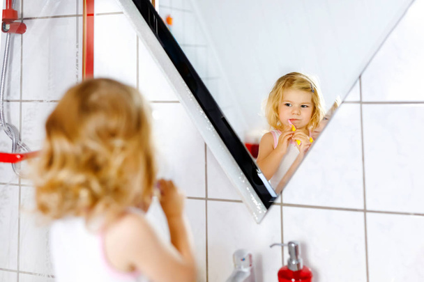 Cute adorable toddler girl holding toothbrush and brushing first teeth in bathroom after sleeping. Gorgeous baby child learning to clean milk tooth. Morning healthy hygiene routine for children - Foto, Bild