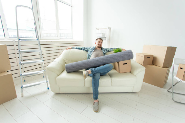 Positieve jonge man zittend op de Bank met zijn dingen in de nieuwe woonkamer na de verhuizing. Concept van de betaalbaarheid van woningen voor jongeren. - Foto, afbeelding