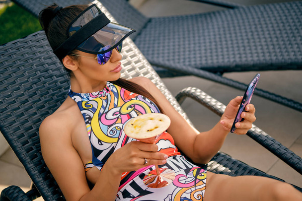 Meisje is poseren leggen op een ligstoel in de buurt van een zwembad en met behulp van haar Smartphone. Gekleed in een kleurrijk badpak, zonneklep en zonnebril. - Foto, afbeelding