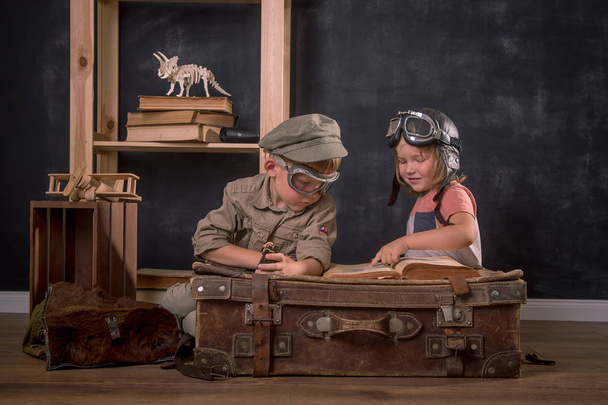 パイロットを演じる子供の男の子と女の子。ビンテージレトロな子供たちは双眼鏡とコンパスを手に古いスーツケースに座ってメガネで肖像画。イノベーション、旅行、調査. - 写真・画像