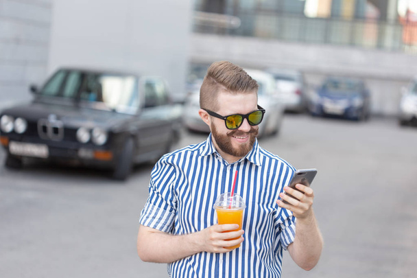 Vista lateral de un joven y elegante hombre hipster con gafas y un bigote y jugo en sus manos charlando en las redes sociales usando un teléfono inteligente e internet inalámbrico en un cálido día de verano
. - Foto, Imagen