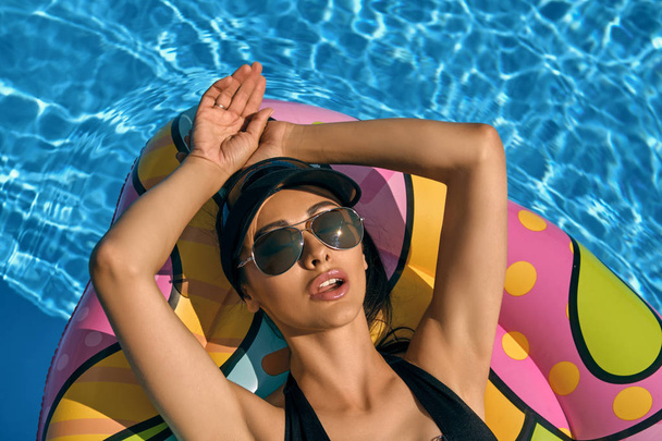Portret kobiety o odpoczynku i stwarzających w basenie na dmuchany materac. Ubrany w czarny strój kąpielowy, osłona przeciwsłoneczna i okulary przeciwsłoneczne. - Zdjęcie, obraz