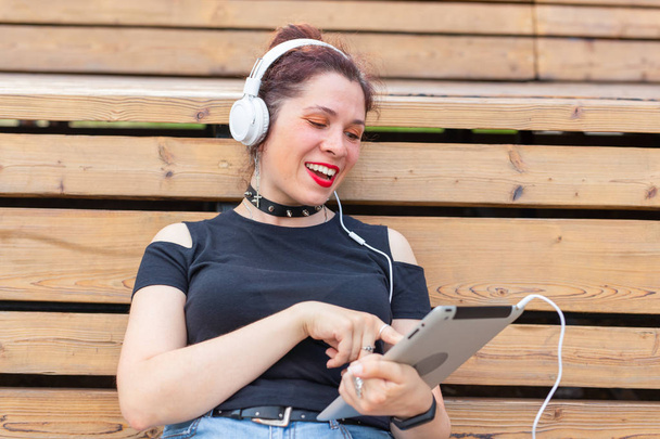 Portret van een mooie stijlvolle jonge vrolijke vrouw in een koptelefoon zittend in het Park op een warme zomerdag. Het concept van vrije tijd en studie in het weekend. - Foto, afbeelding