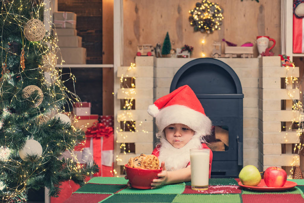 Μπισκότα για τα παιδιά του Αϊ-Βασίλη. Ευτυχισμένος Άγιος Βασίλης-χαριτωμένο αγόρι παιδί τρώει ένα μπισκότο και πίνοντας ποτήρι γάλα στο σπίτι Χριστούγεννα εσωτερικό. - Φωτογραφία, εικόνα