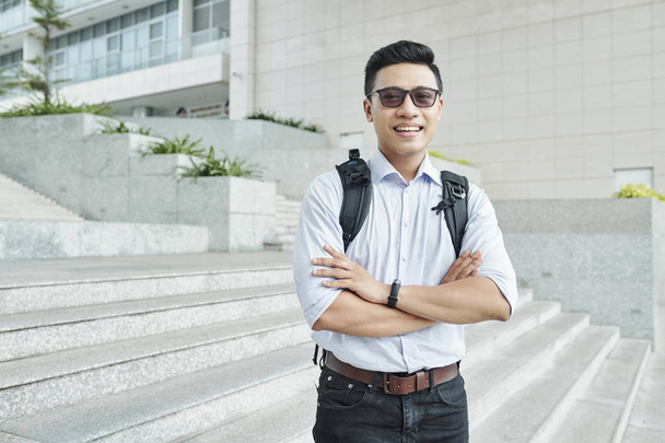 fröhlicher vietnamesischer Student in Sonnenbrille, der mit verschränkten Armen auf Stufen vor der Universität steht - Foto, Bild