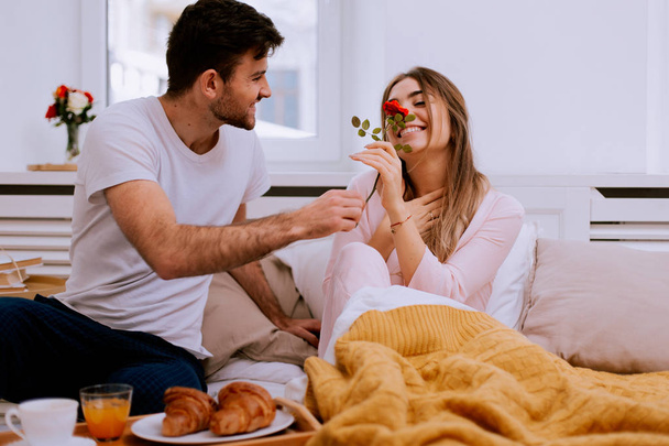romantisches Frühstück im Bett Mann bereitet für seine Frau, sie war sehr glücklich, Mann bringen eine rote Rose für sie riecht sie die Blume und war sehr beeindruckt - Foto, Bild