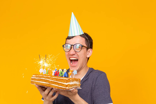 Funny positive guy in glasses holds in his hands a homemade cake with the inscription happy birthday posing on a yellow background. Concept of holidays and anniversaries. - Photo, Image