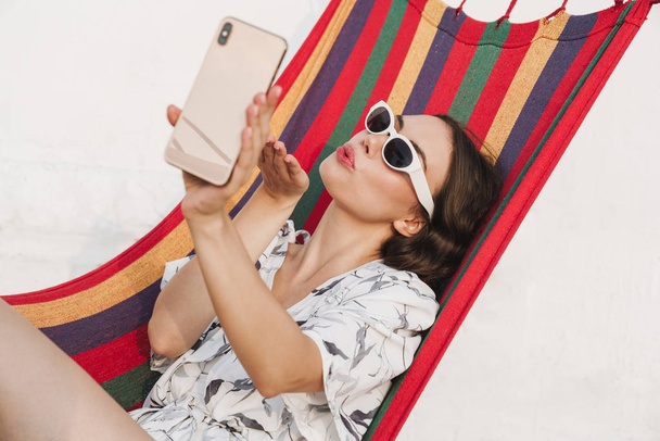 Young beautiful woman at the beach posing on a hammock take a selfie by mobile phone. - Foto, Imagem