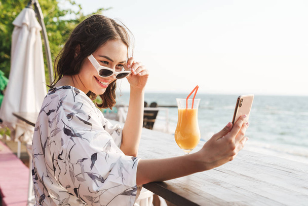Cute happy young beautiful woman at the beach drinking juice cocktail in cafe wearing sunglasses take a selfie by phone. - Фото, изображение