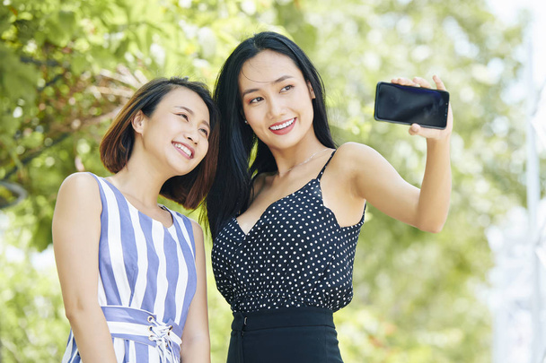 2人の笑顔のアジアの女性が立って、街の屋外で携帯電話で一緒に自分撮りポートレートを作る - 写真・画像
