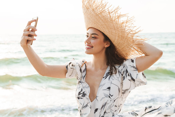 Pleased happy young beautiful woman at the beach walking in a beautiful sunny morning take a selfie by mobile phone. - Photo, Image