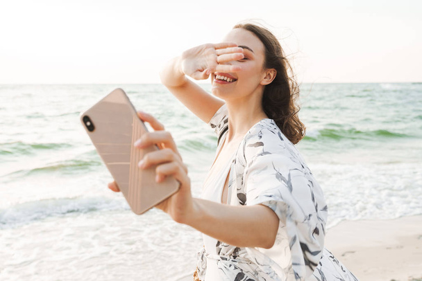 Pleased happy young beautiful woman at the beach walking in a beautiful sunny morning take a selfie by mobile phone. - Φωτογραφία, εικόνα