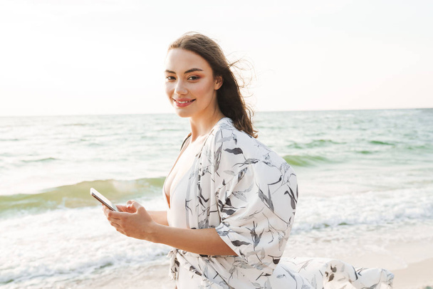 Sonriendo feliz feliz joven hermosa mujer en la playa caminando en una hermosa mañana soleada utilizando el teléfono móvil
. - Foto, Imagen