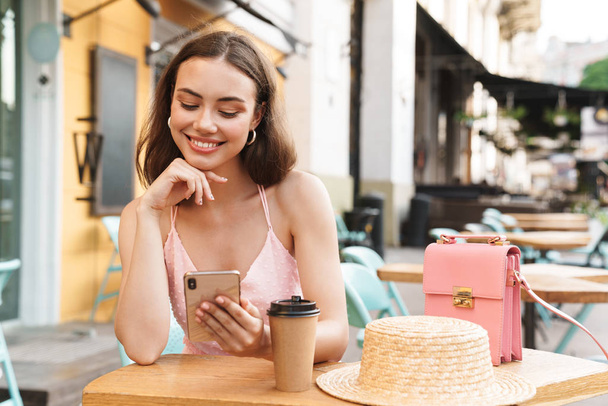 Wizerunek zadowolony brunetka kobieta uśmiechając się i trzymając telefon komórkowy siedząc w letniej kawiarni ulicy - Zdjęcie, obraz
