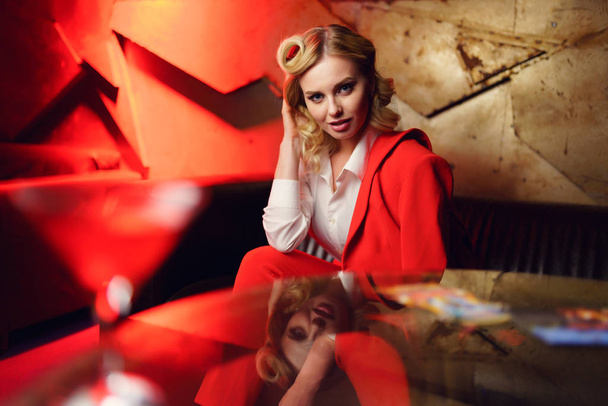 Photo of trendy blonde in red jacket sitting on sofa at table - Fotó, kép