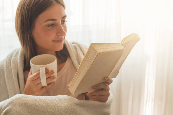 Το όμορφο κορίτσι διαβάζει ένα βιβλίο με ένα φλιτζάνι ζεστό τσάι. Χειμερινή ώρα φθινοπώρου. - Φωτογραφία, εικόνα