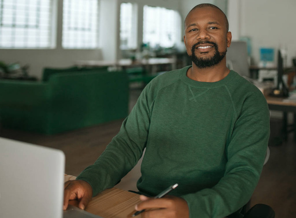 Ritratto di un sorridente designer afroamericano che lavora su un computer portatile seduto alla scrivania del suo ufficio nel tardo pomeriggio
 - Foto, immagini