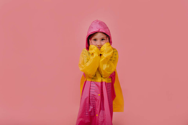 Porträt eines hübschen kleinen Mädchens in gelbem Regenmantel, das sich auf Regen vorbereitet, isoliert auf rosa Hintergrund - Foto, Bild