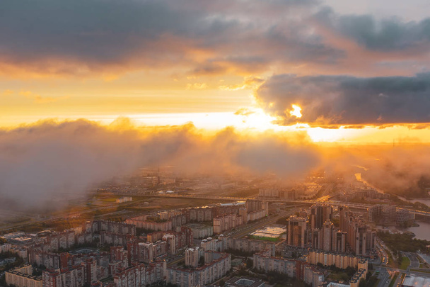 都市景観、都市の住宅地の上に低空飛行雲と空中トップビュー、地平線と太陽の夜明けのビュー. - 写真・画像