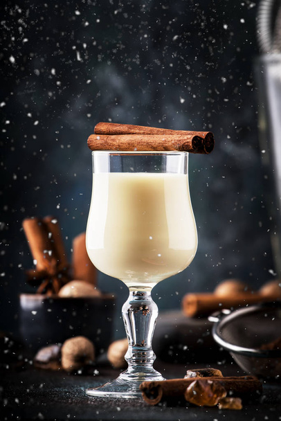 Eggnog cocktail hot winter or autumn drink with milk, eggs and dark rum, sprinkled with cinnamon and nutmeg in shot glass on blue dark background, copy space - Foto, afbeelding