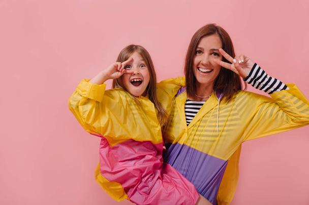 Close up portrait of pretty woman wearing bright raincoat holding her little charming daughter and showing peace sign  - Fotoğraf, Görsel
