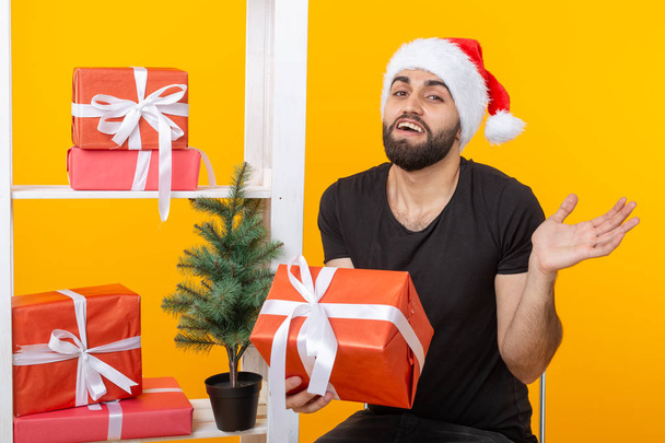 Noel Baba şapkalı genç yakışıklı adam bir Noel ağacının yanında tebrik hediyeleri tutuyor. Noel ve yeni yıl tatili kavramı. - Fotoğraf, Görsel