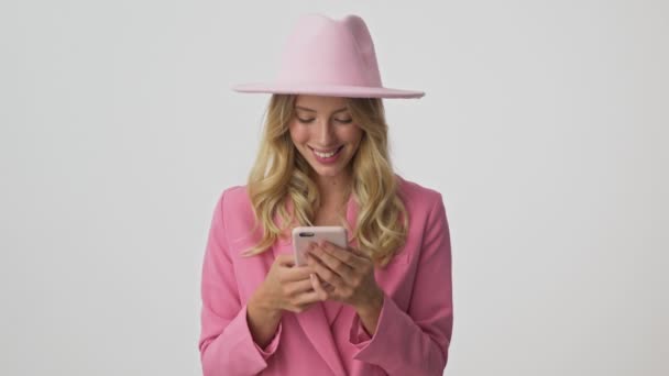 Cheerful young blonde woman in pink jacket and hat smiling and laughing while using smartphone over gray background isolated - Metraje, vídeo