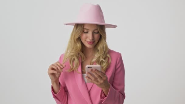 Pensive young blonde woman in pink jacket and hat smiling and playing with her hair while using smartphone over gray background isolated - Video