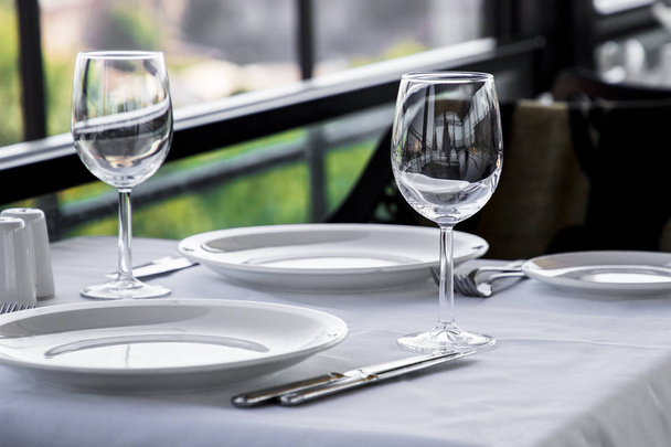 Table setting in restaurant terrace with green background. Empty wine glasses, plates, forks, knifes on the tablecloth. Elegant restaurant interior. - Photo, Image