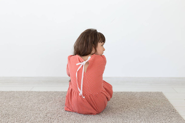 Vista trasera de una niña pequeña con un vestido de lunares rojo sentado en el suelo sobre un fondo blanco. El concepto de productos para niños
. - Foto, Imagen